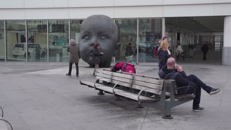 People-Taking-Pictures-At-Day-and-Night-Sculpture-By-Artist-Antonio-Lopez,-A-Giant-Baby-Head-Sculpture-In-Atocha-Railway-Station-In-Madrid,-Spain