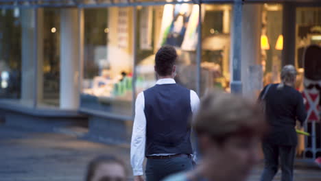 Un-Joven-Elegante-Con-Una-Sencilla-Mascarilla-Protectora-Verde-Covid-19,-Un-Chaleco-Negro,-Camisa-Blanca-Y-Jeans-Cruzando-La-Calle,-De-Espaldas-A-La-Cámara,-Una-Tienda-De-Alta-Gama-Al-Fondo,-4k