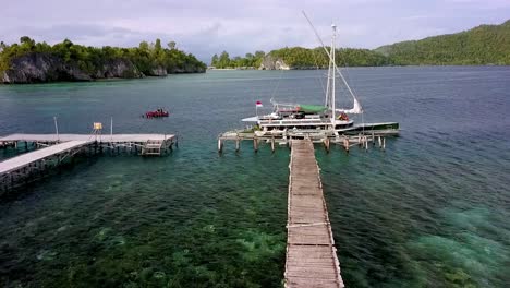 Boat-with-passengers-at-a-damaged-docking-pier-displaying-the-Indonesian-flag-while-the-dinghy-tours-snorkelers,-Aerial-dolly-in-rising-shot