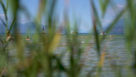 Plano-General-De-Personas-Nadando-En-El-Lago-Con-Botes-Que-Pasan,-Disparados-A-Través-De-Juncos-En-La-Playa-De-Jamaica,-Sirmione,-Lago-Garda,-Lago-De-Garda,-Italia