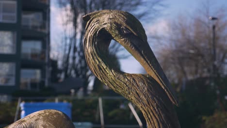 Eine-Nahaufnahme-Einer-Blaureiher-Skulptur-In-Der-Geschäftigen-Stadtlandschaft-Mit-Einem-Verschwommenen-Hintergrund-Von-Gebäuden-Und-Einem-Baum-Mit-Blauem-Himmel