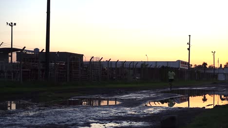 Un-Corredor-Solitario-Al-Atardecer-En-Una-Vía-De-Servicio-Inundada-Junto-A-Una-Fábrica