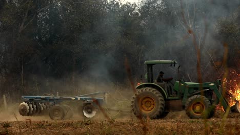 Traktor-Fährt-über-Ein-Feld-Mit-Einem-Brennenden-Buschwerk-Im-Hintergrund