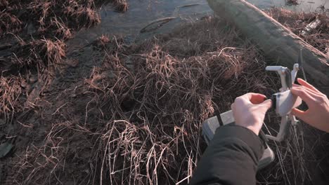 Blick-über-Die-Schulter-Einer-Person,-Die-Den-Nd-Filter-An-Ihrem-Kleinen-DJI-Mini-Drohnen-UAV-Flugzeug-Anpasst,-Mit-Gras-Sand-Schlamm-Logs-Im-Hintergrund