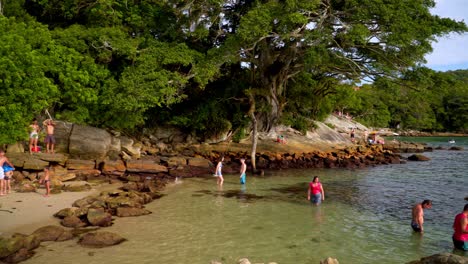 Pan-left-of-people-relaxing-near-the-sea-in-a-rocky-shore-surrounded-by-woods-in-Praia-da-Sepultura,-Brazil