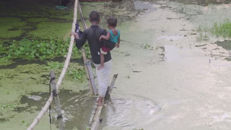 Niño-Cargando-A-Un-Niño-En-Agua-Inundada-En-La-Parte-Superior-De-Un-Puente-De-Bambú