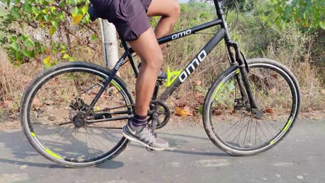 Un-Joven-Ciclista-Indio-Moviéndose-Rápido-En-Bicicleta-En-Medio-De-La-Carretera-Rural-Con-Plantas-Y-Manglares-En-El-Fondo,-Deportes-Y-Fitness,-Entrenamiento,-Ejercicio,-Concepto-De-Entrenamiento-Para-Jóvenes