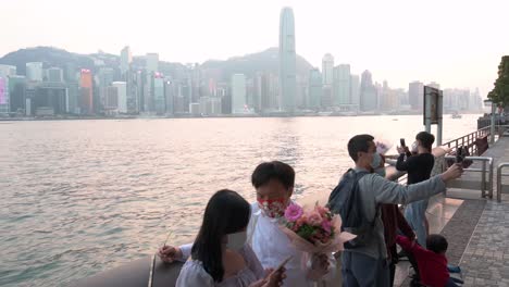 Una-Pareja-Se-Toma-Una-Selfie-Con-Una-Boutique-De-Rosas-Mientras-La-Gente-Disfruta-De-La-Vista-Frente-Al-Mar-Del-Puerto-Victoria-Del-Horizonte-De-La-Isla-De-Hong-Kong-Mientras-Se-Pone-El-Atardecer