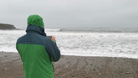 Hombre-De-Pie-En-La-Playa-En-Un-Clima-Tormentoso-Tomando-Fotos-Con-Olas-Rompiendo-En-La-Orilla