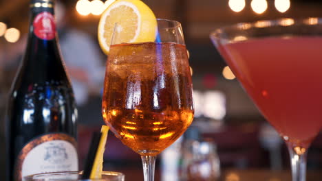 La-Bebida-Aperol-Spritz-Se-Encuentra-Entre-Una-Botella-De-Vino-Y-Un-Cosmo-Rodeado-De-Luces-Cálidas-En-Un-Bar-Oscuro,-Deslizador-4k