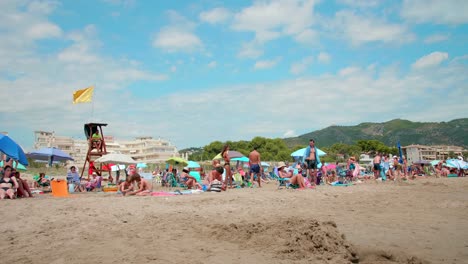 Yellow-Flag-On-Lifeguard-Post-At-The-Beach-In-Alcossebre,-Spain-With-Tourists-Relaxing-In-Summer