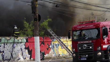 Bombero-Sofoca-Un-Gran-Incendio-Urbano-Con-Humo-Desde-Lo-Alto-De-Un-Muro-De-Hormigón,-En-La-Avenida-Tereza-Cristina,-Distrito-De-Ipiranga