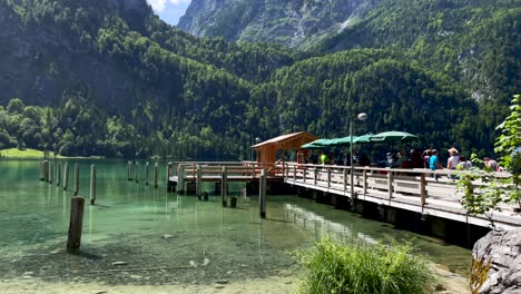 Schwenkaufnahme-Von-Touristen,-Die-Am-Holzsteg-Des-Hafens-Eines-Elektroboots-Auf-Eine-Kreuzfahrt-Auf-Dem-Königssee-In-Deutschland-Warten---Wunderschöne-Berglandschaft-Im-Sommer