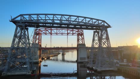 Historic-monument-Puente-Transbordador-Nicolas-Avellaneda-bridge-at-sunrise-at-La-Boca-neighborhood-with-a-small-boat-rowing-on-matanza-river,-Buenos-Aires,-Argentina,-aerial-front-view-pan-shot