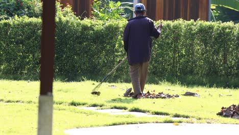 4K-Male-Gardener-Raking-the-Leaves-from-a-Grass-Lawn-Garden-in-Thailand