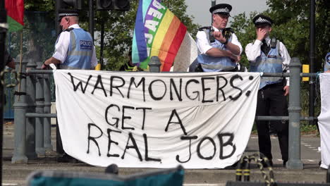 Verbindungsbeamte-Der-Stadtpolizei-Stehen-Bei-Einer-Protestaktion-Gegen-Die-Dsei-Waffenmesse-Hinter-Einem-Weißen-Banner-Mit-Schwarzer-Aufschrift-Mit-Der-Aufschrift-„Kriegstreiber,-Sucht-Euch-Einen-Richtigen-Job“.