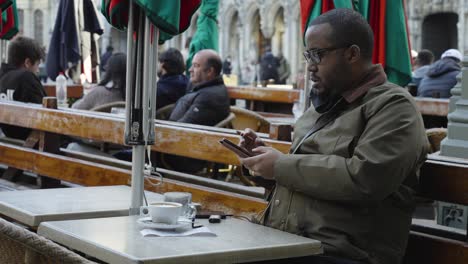 Hombre-De-Piel-Oscura-De-Mediana-Edad-Desplazándose-Por-El-Teléfono-Inteligente-Mientras-Bebe-Café-Y-Fuma-Un-Cigarro-En-Una-Terraza-De-La-Ciudad