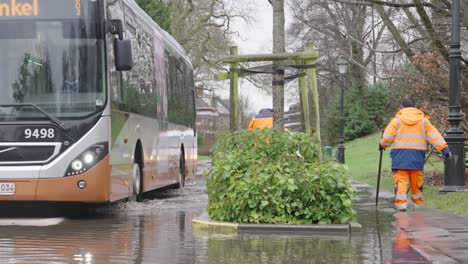 Conductor-De-Autobús-Afroamericano-En-La-Calle-Inundada-Pasando-Trabajadores-Municipales-Reparando-El-Alcantarillado-Después-De-Fuertes-Lluvias-En-El-Woluwepark-En-Bruselas,-Bélgica