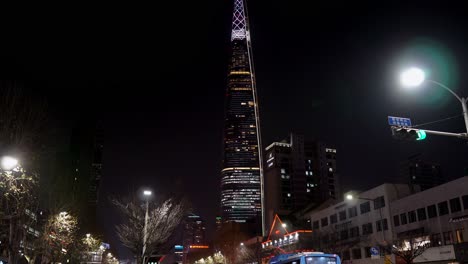 Inclínate-En-La-Torre-Lotte-World-Por-La-Noche-Mientras-Las-Personas-Con-Máscaras-Protectoras-Esperan-El-Autobús-En-La-Parada-De-Autobús