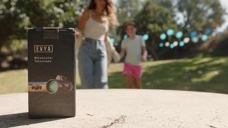 product-shot-of-a-telescope-box-while-in-the-background-a-mother-and-daughter-approaching