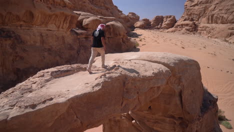 Hombre-Caminando-Sobre-Un-Puente-De-Arco-Natural-De-Arenisca-Con-Un-Impresionante-Mirador-Sobre-El-Paisaje-Desértico-De-Wadi-Rum,-Jordania