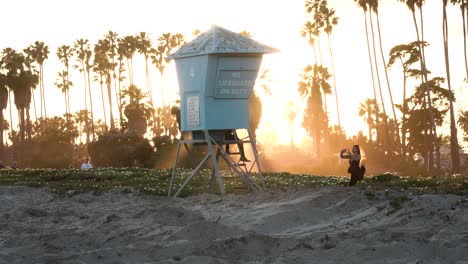 Two-friends-taking-pictures-on-lifeguard-tower-in-East-beach-during-sunset-with-palm-trees,-Handheld-shot