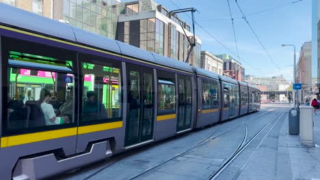 The-Dublin-tram-arrives-at-the-Abbey-Street-stop,-with-many-people-walking-through-the-streets-on-a-sunny-day