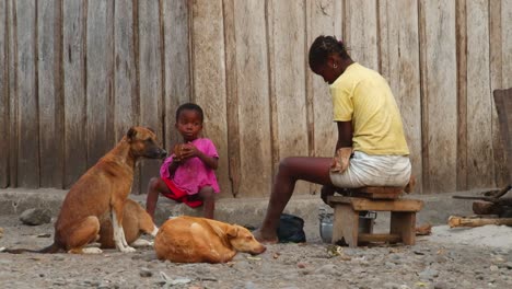 Un-Niño-Se-Sienta-Al-Lado-De-Tres-Perros-Y-Come-Al-Lado-De-Su-Madre-Africana-En-El-Suelo-Afuera-Mientras-Ella-Remueve-La-Olla---Cierra