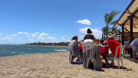 Familia-Sentada-En-La-Playa-Y-Comiendo-En-Un-Restaurante-Con-Vista-Al-Mar-En-Un-Día-Ventoso-En-Marbella-España,-Clima-Soleado-Y-Cielo-Azul-Con-Palmeras,-Tiro-De-4k