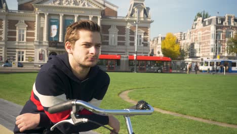 Hübscher-Junger-Mann,-Der-An-Einem-Sonnigen-Tag-Auf-Einem-Fahrrad-In-Der-Nähe-Der-Konzerthalle-In-Amsterdam-Steht