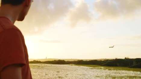 Joven-Modelo-Masculino-Está-Posando-En-La-Luz-Dorada-Del-Atardecer-Mientras-El-Avión-Aterriza-En-Un-Fondo