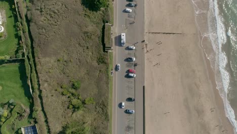 Luftaufnahme-Von-Oben-Nach-Unten-über-Ein-Luxushaus-Auf-Einer-Klippe-Und-Eine-Strandstraße-Mit-Geparkten-Autos-Und-Verkehr-In-Beide-Richtungen