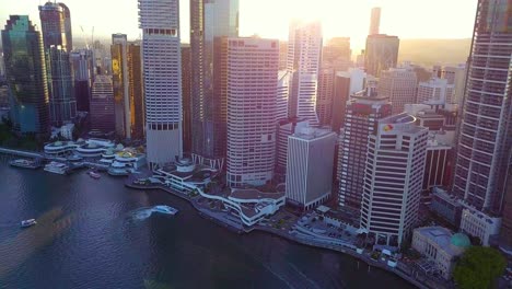 Vista-Aérea-De-Una-Ciudad-Moderna-Con-Edificios-Altos-Y-Ferry-Público-Junto-Al-Río-Al-Atardecer