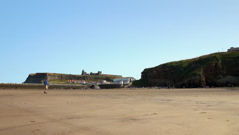 Cámara-Estática-De-4k-Que-Muestra-A-Un-Hombre-Caminando-Solo-Por-Una-Playa-Tranquila-Con-La-Abadía-De-Whitby-En-Una-Colina-Al-Fondo