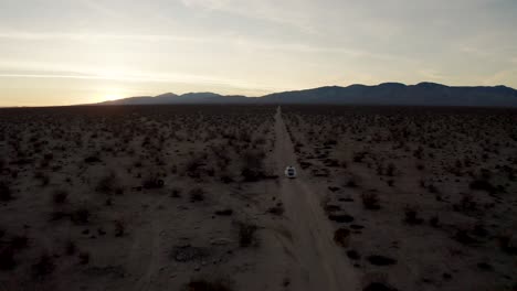 Toma-Aérea-De-Un-Automóvil-Blanco-Conduciendo-Por-Un-Sendero-A-Través-De-La-Llanura-Del-Desierto-De-Mojave