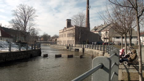 Ein-Elternteil-Und-Ein-Kind-Im-Kinderwagen-Sitzen-Auf-Einer-Bank-Mit-Blick-Auf-Den-Fluss-Chrudimka-In-Hlinkso,-Tschechische-Republik