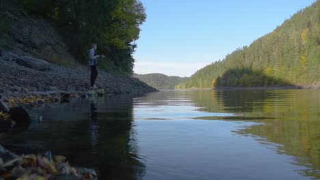 Young-man-is-standing-at-the-shore-of-the-local-lake-with-mountains-in-the-background-while-fishing-with-a-fishing-rod-on-a-sunny-day