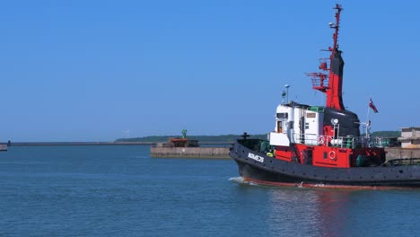 Black-and-red-colored-harbour-tug-leaving-Port-of-Liepaja-in-hot-sunny-day,-medium-shot