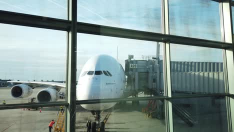 View-through-airport-terminal-window-of-huge-aircraft-waiting-at-gate-as-ground-crew-prepares-for-customers-to-disembark-and-board-for-return-flight