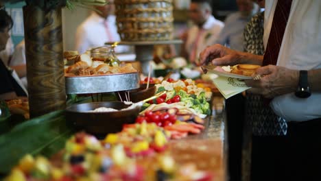 Smooth-shot-of-a-line-of-people-at-a-crowded-event-going-through-a-food-line-and-grabbing-a-variety-of-food-including-meats,-cheeses,-bread,-fruits,-and-vegetables