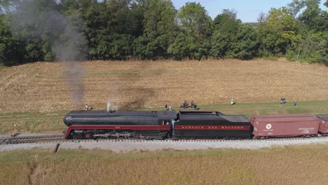 Vista-Aérea-De-Un-Motor-De-Vapor-Antiguo-Que-Se-Aleja-De-La-Estación-Con-Vagones-De-Carga-A-Través-De-Tierras-Agrícolas-Amish-En-Un-Día-Soleado-De-Otoño-Con-Una-Vista-De-Distancia-Vista-Por-Un-Dron