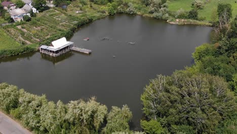 Aerial-View-of-Rugged-Shack-on-a-River-With-a-Dock-and-Houses-in-Background-on-a-Sunny-Day