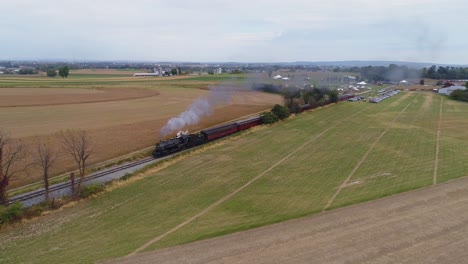 Luftaufnahme-Einer-Restaurierten-Dampfmaschine,-Die-Dampf-Und-Rauch-Bläst,-Während-Sie-An-Einem-Sonnigen-Tag-Antike-Personenkraftwagen-Mit-Blick-Auf-Ackerland-Zieht