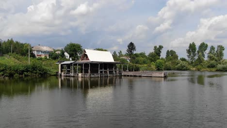 Low-Aerial-View-of-Rugged-Shack-on-a-River-With-a-Dock-and-Houses-in-Background-on-a-Sunny-Day