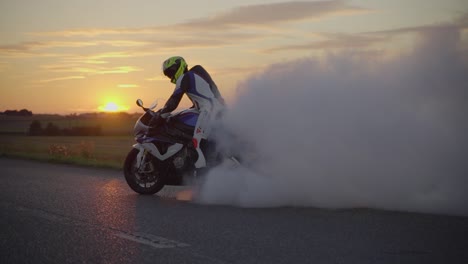 Toma-Media-Ancha-De-Un-Motociclista-Con-Traje-De-Cuerpo-Entero-Y-Casco-Realizando-Un-Agotamiento-Con-Una-Superbike-Blanca-Durante-La-Puesta-De-Sol.