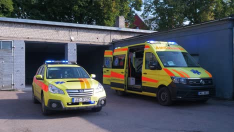 Dos-Vehículos-De-Ambulancia-Paramédicos-Amarillos-Estacionados-Fuera-Del-Garaje-De-Ladrillo-Blanco,-Luces-De-Advertencia-Azules-Intermitentes,-Día-Soleado,-Tiro-Ancho-De-Mano