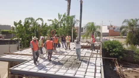 Vista-Aérea-De-Los-Trabajadores-De-La-Construcción-Que-Trabajan-En-El-Techo-Vertiendo-Cemento-Para-Construir-Una-Estructura-De-Techo-O-Piso-Usando-Chalecos-Naranjas