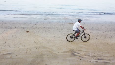 Un-Joven-Indio-Marrón-Montando-Bicicleta-Temprano-En-La-Mañana-En-La-Orilla-De-La-Playa-Con-Un-Dron-Disparó-Un-Fondo-De-Video-Con-Vista-Aérea,-Un-Joven-Indio-Haciendo-Ejercicio-En-La-Orilla-De-La-Playa