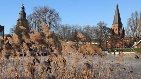 Eislaufende-Menschen-Auf-Dem-Kanal-Mit-Schilf-Im-Vordergrund-Und-Historischen-Mittelalterlichen-Gebäuden-Im-Hintergrund