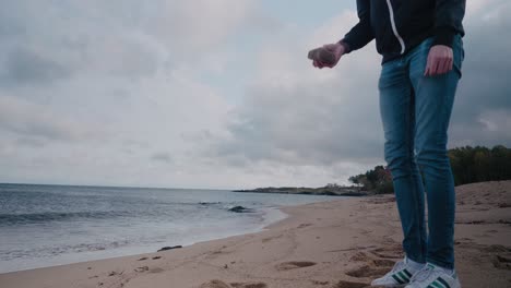 Young-Tall-Guy-With-Jacket-and-Cap-Picking-and-Choosing-Stones-To-Through-Into-the-Sea---Static-Wide-Shot